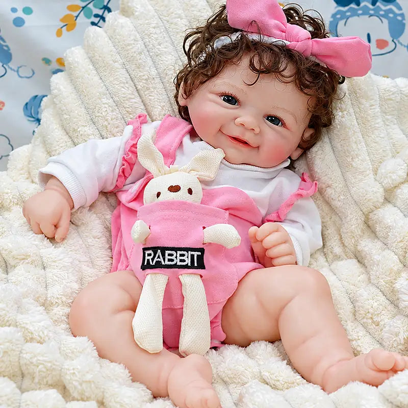 Realistic silicone baby Layla with curly brown hair, bright eyes, rosy cheeks, and a sweet smile, dressed in pink and holding a soft bunny toy.