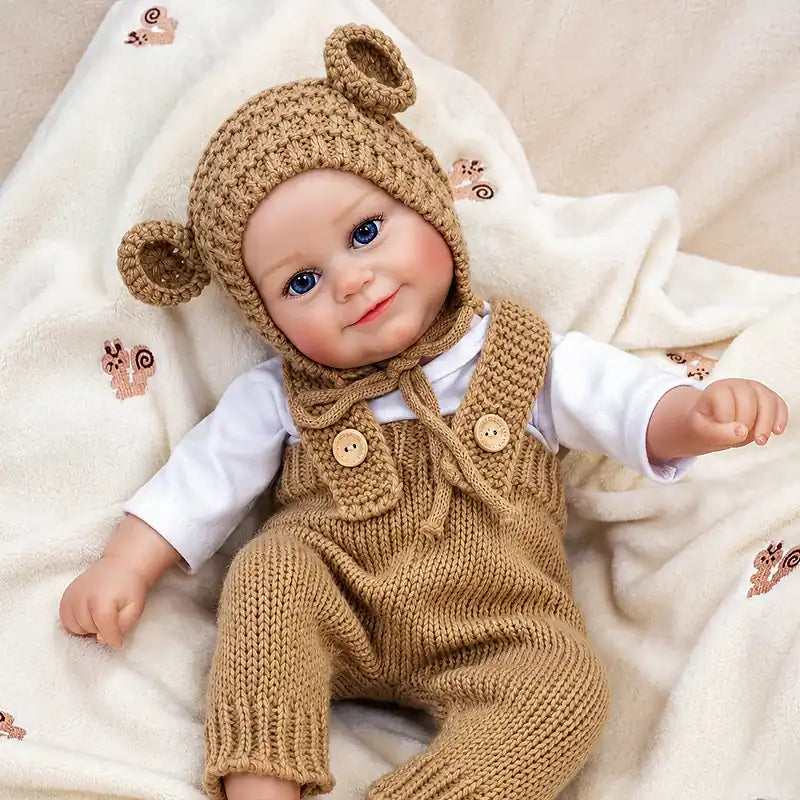 Reborn doll wearing a crocheted bear-ear hat and buttoned knit overalls over a white shirt.