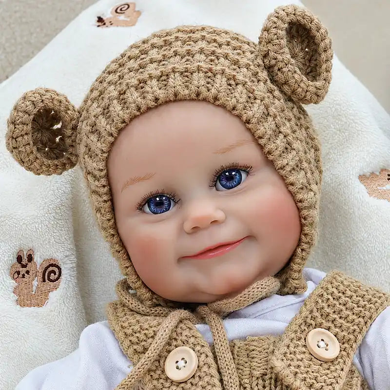 Close-up of a smiling reborn doll with blue eyes, rosy cheeks, and a caramel knit bear hat.