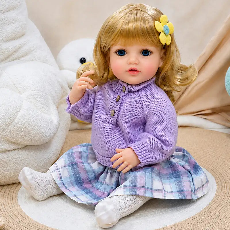 Lifelike reborn baby doll with blonde curly hair, bright blue eyes, lavender knit sweater, plaid skirt, and teddy bear.