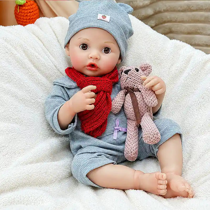 Realistic reborn doll with a playful seated pose, soft blue romper, red scarf, bear hat, tiny toes, and teddy bear accessory for collectors and gift giving.