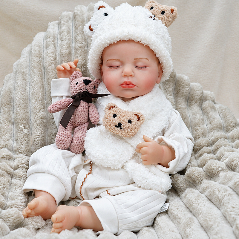 Collectible realistic reborn baby Ollie with lifelike closed eyes, rosy cheeks, glossy lips, tiny toes, a textured white outfit, and a knitted teddy bear companion.