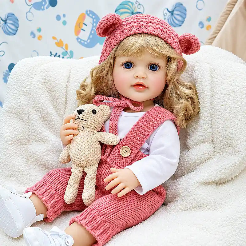 Realistic reborn doll Isabella wearing a pink knitted romper and bear-ear hat, holding a tiny teddy bear, perfect for cuddles, pretend play, and nursery display.