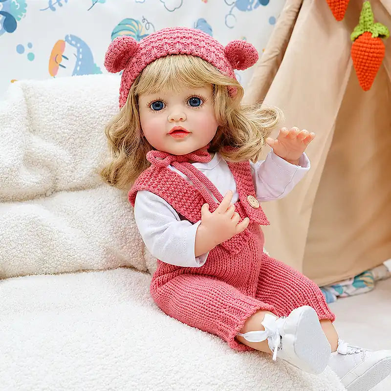 Lifelike doll Isabella with golden curly hair, bright blue eyes, rosy cheeks, glossy lips, and delicate fingers, dressed in soft textured knitwear for a cozy handcrafted look.