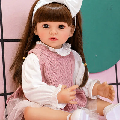 Brunette reborn girl doll with straight bangs, side braids, large brown eyes, and a charming pink-and-white layered dress.