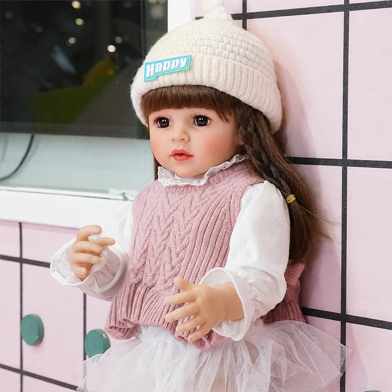 Fashion reborn girl doll wearing a pink cable-knit dress, white ruffle blouse, and soft tulle skirt with long brunette hair.