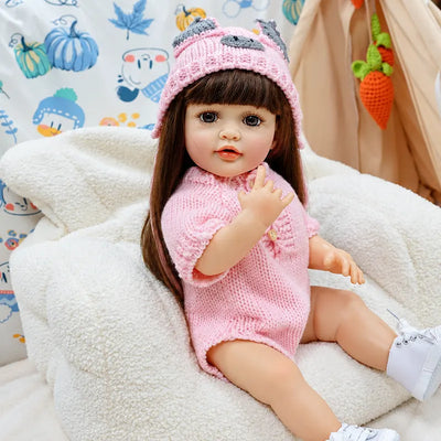 Lifelike reborn doll with soft pink skin, lifelike brown eyes, long brunette hair, and a detailed knitted outfit, photographed in a warm bedroom nursery scene.