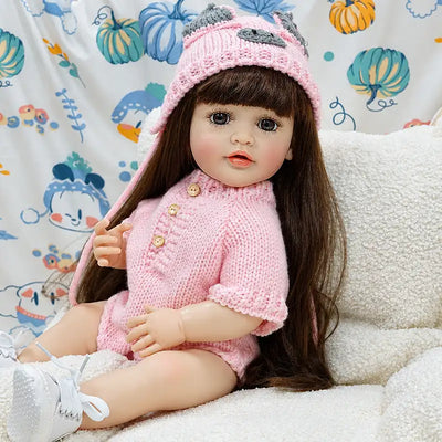 Highly detailed reborn doll with natural facial coloring, bright lifelike eyes, delicate fingers, long brown hair, and carefully textured pink knit clothing.