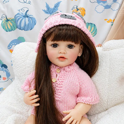 Realistic reborn baby doll wearing a handmade pink knitted romper and matching hat, seated on a cozy bed with long brown hair, glossy eyes, and delicate facial details.