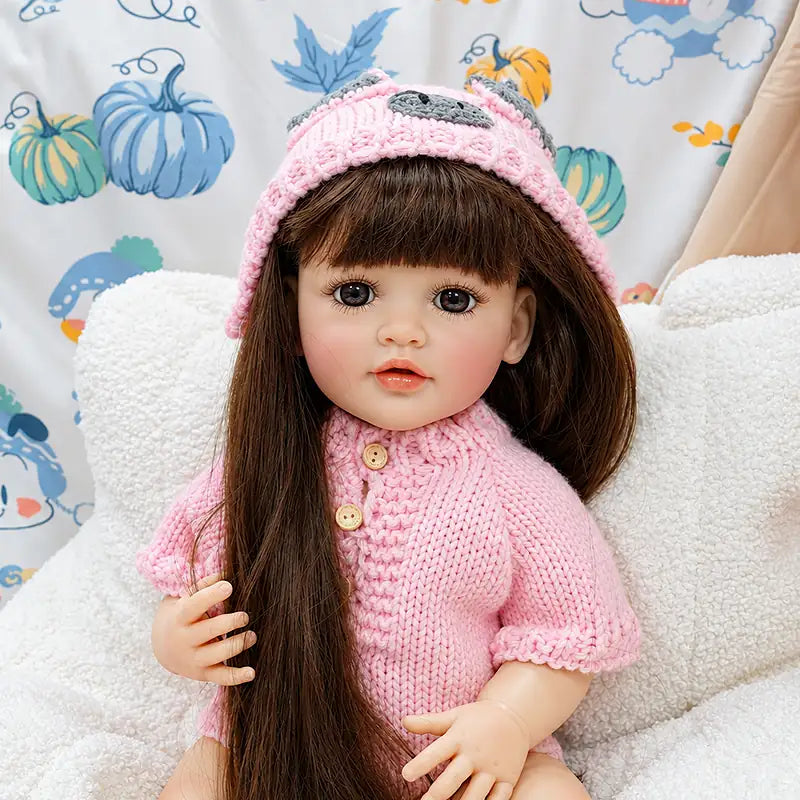 Realistic reborn baby doll wearing a handmade pink knitted romper and matching hat, seated on a cozy bed with long brown hair, glossy eyes, and delicate facial details.