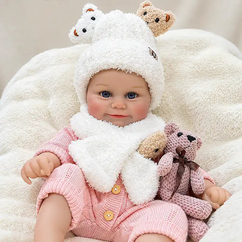 Smiling reborn doll with bare little feet, a pink knit outfit, and a teddy bear scarf.