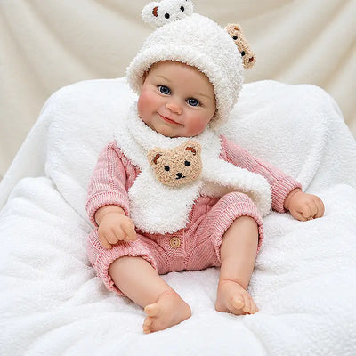 Soft vinyl reborn doll sitting on a white blanket, dressed for cozy pretend play.