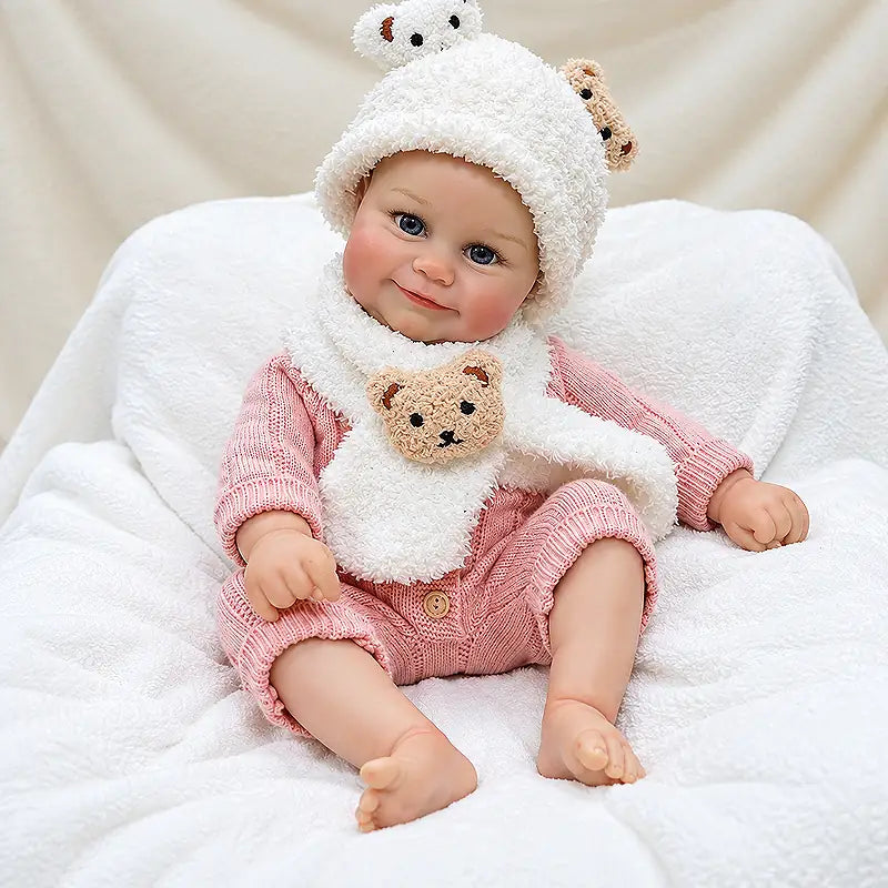 Soft vinyl reborn doll sitting on a white blanket, dressed for cozy pretend play.