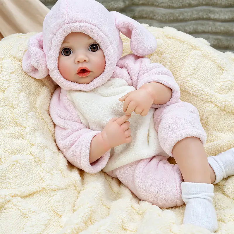 Realistic baby doll Ella with a bottle-feeding function, soft-looking limbs, delicate facial features, a pink bunny hood, and a cuddly teddy bear accessory.