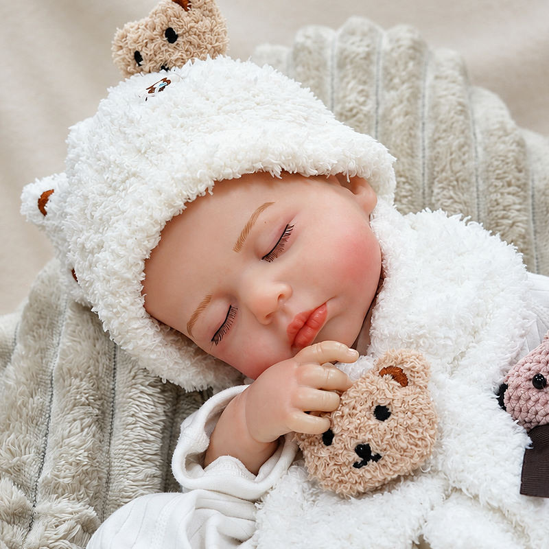 Realistic baby doll Ollie sleeping on a soft blanket while holding a knitted teddy bear, perfect for nursery décor, newborn photography, and pretend bedtime play.