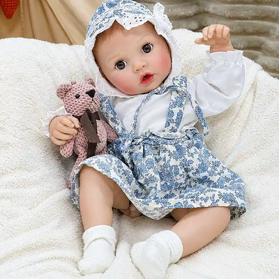 Realistic reborn baby Ellis wearing a blue floral outfit and bonnet, with bright brown eyes, delicate hands, rosy cheeks, and a bottle-feeding feature.