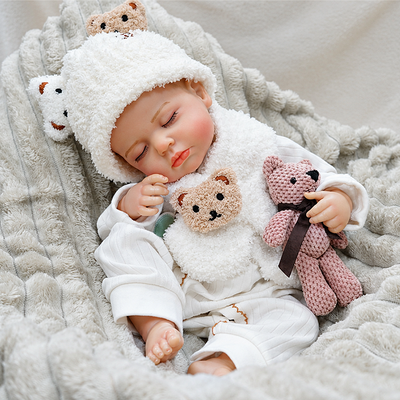 Realistic reborn baby Ollie wearing a fluffy white bear outfit, with closed eyes, rosy cheeks, tiny toes, and a cuddly teddy bear accessory.
