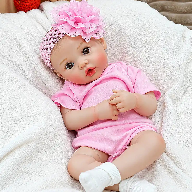 Realistic reborn feeding baby doll Maisie wearing a soft pink romper and oversized flower headband, with bright eyes, rosy cheeks, glossy lips, and delicate hands.