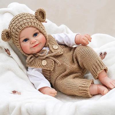 Reborn baby doll in a matching bear hat and knit overalls, posed on a soft blanket.