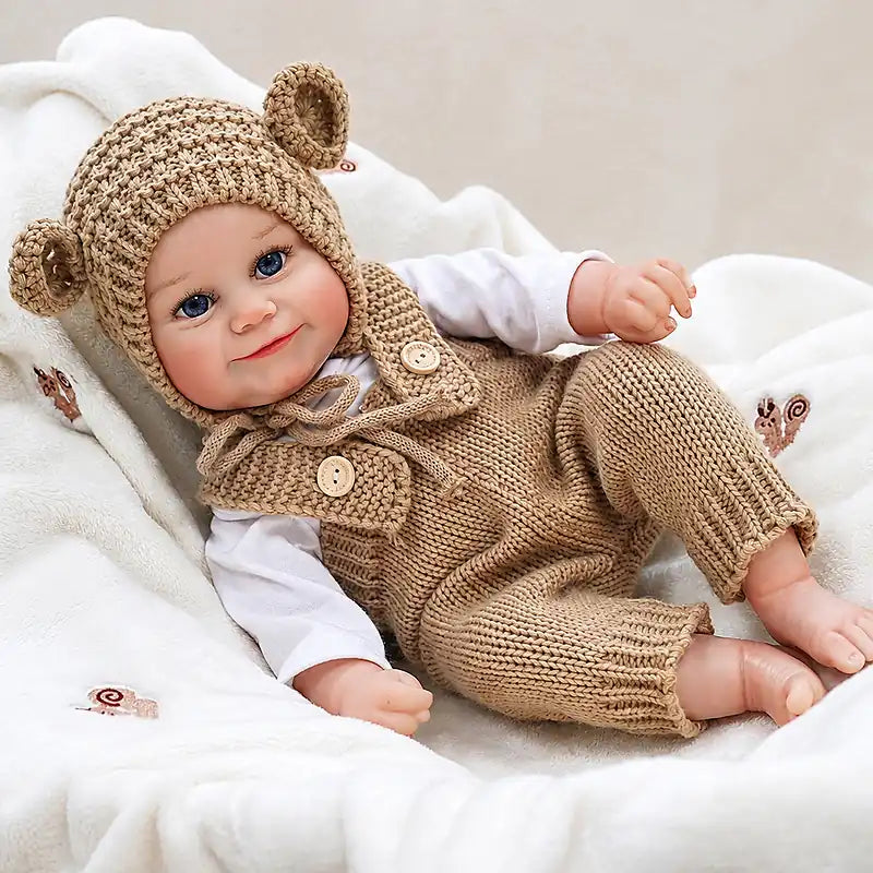 Reborn baby doll in a matching bear hat and knit overalls, posed on a soft blanket.