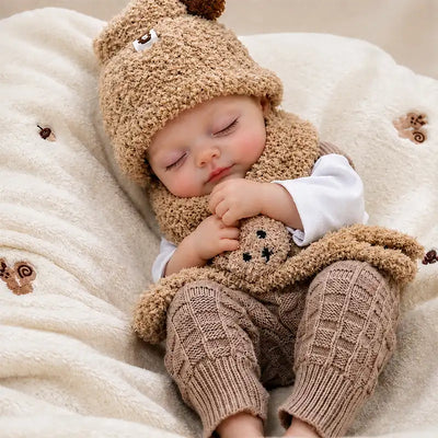 Realistic sleeping reborn baby doll with lifelike facial details, rosy cheeks, brown knitted clothes, bear hat, scarf, and teddy bear in a cozy nursery setting.