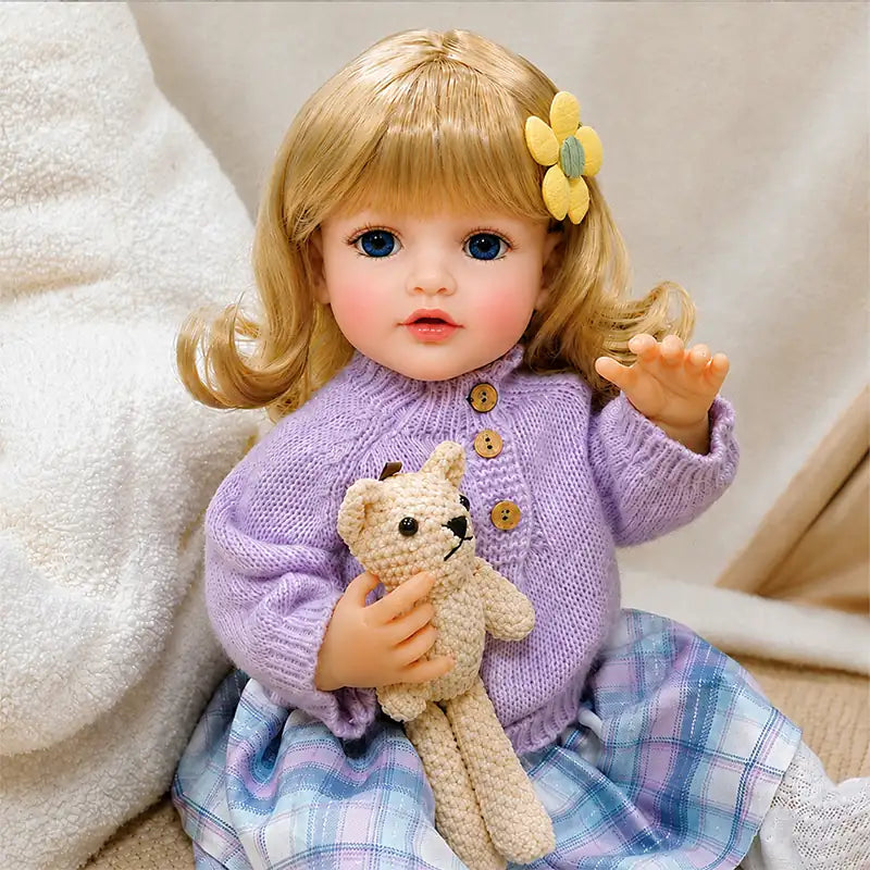 Blonde reborn baby doll sitting in a cozy nursery setting while holding a teddy bear, perfect for pretend play and quiet cuddle time.