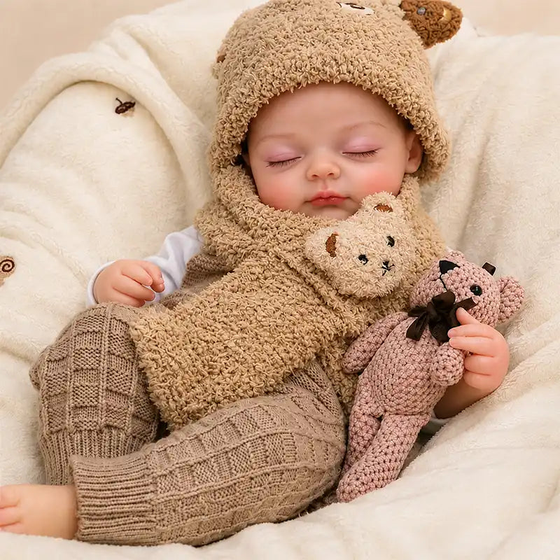 Adorable sleeping reborn baby doll wearing a fluffy bear hat and scarf, holding a matching teddy bear for an extra cozy and charming look.