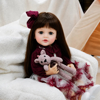 Long-haired realistic reborn girl doll with fashionable clothing, detailed hands, expressive eyes and an adorable sitting pose for play, photography or display.
