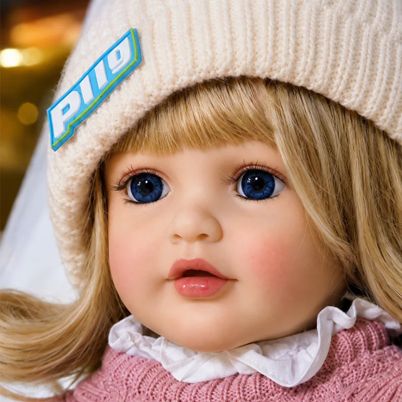 Lifelike blonde reborn toddler doll with blue eyes, rosy cheeks, realistic details, pink knitted clothing, and cream beanie for collectors and pretend play.