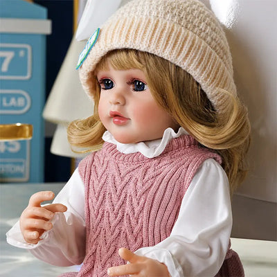 Cozy reborn toddler doll sitting in a relaxed home setting, dressed in soft pink knitwear for a charming everyday childhood-inspired scene.