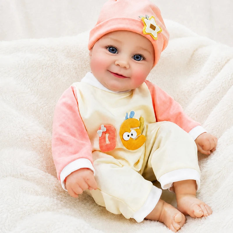 Smiling reborn baby doll wearing a peach hat with star detail and a cream romper, featuring blue eyes, chubby cheeks, tiny fingers, and bare baby feet.