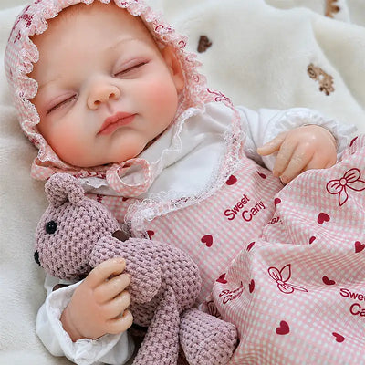 Sleeping reborn baby doll wearing a pink heart-print gingham dress and matching ruffled bonnet, with tiny fingers, realistic proportions, and charming baby details.